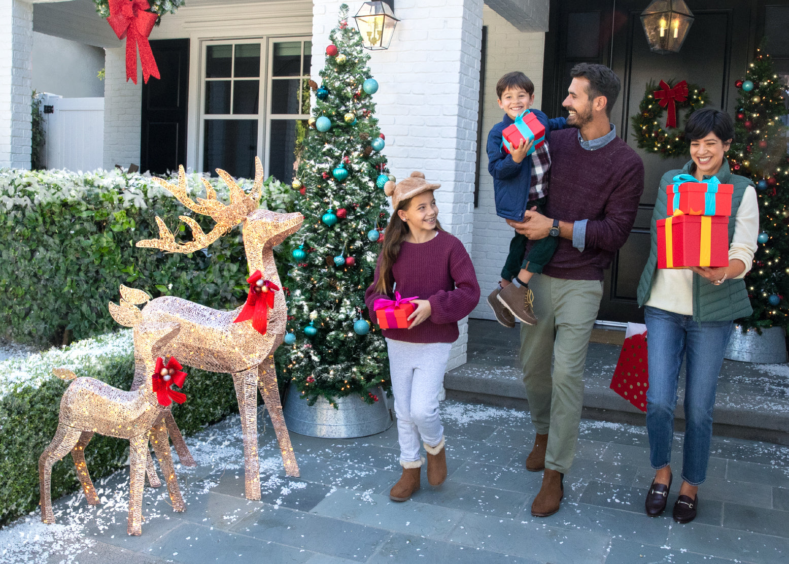 A family walking outside of their house carrying presents. There is a mom, dad, a pre-teen girl and a young boy. Two standing reindeer decorations are on the path.